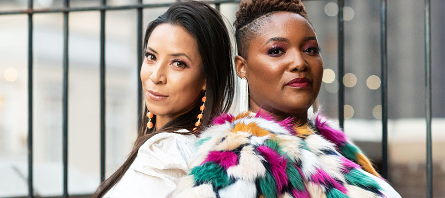 Two women standing back to back in front of a fence with no fear, smizing